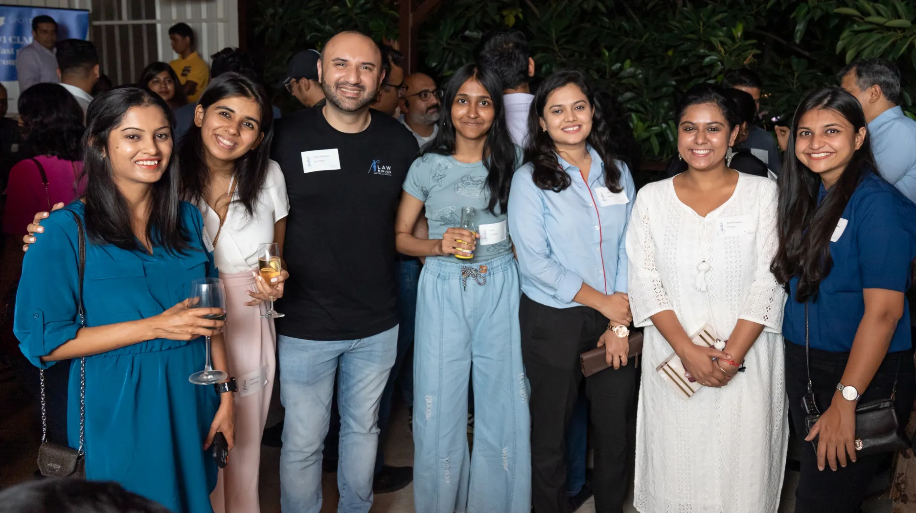 Group of six people smiling and posing at an outdoor event, holding drinks and wearing name tags.